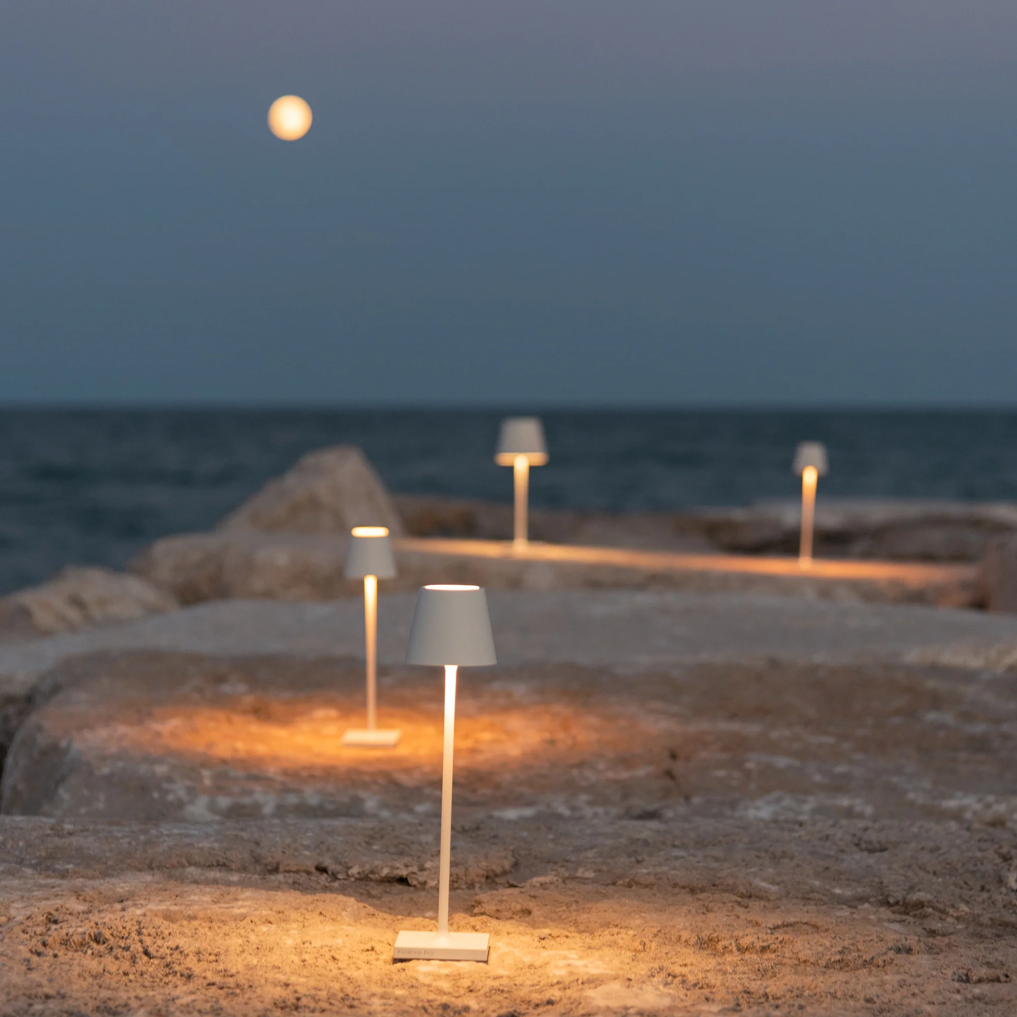 The white Poldina Pro Micro Cordless Lamp from Zafferano illuminating an outdoor path beside a lake.