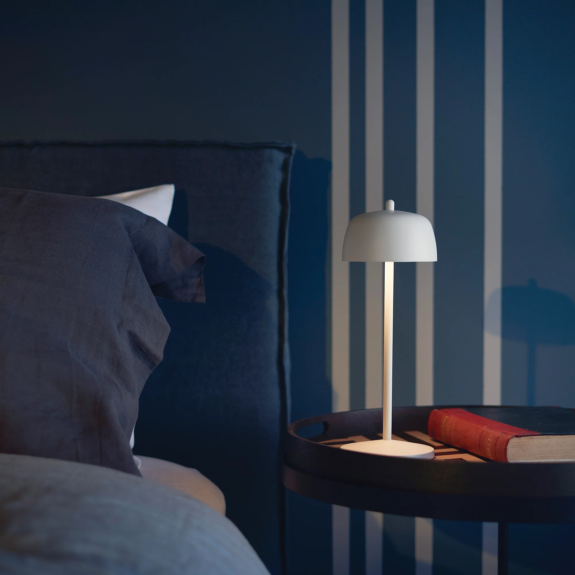 The matte white Theta Pro Cordless Lamp from Zafferano in a bedroom on a bedside table.