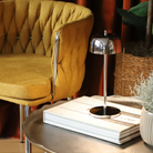 The polished chrome Theta Pro Cordless Lamp from Zafferano on a table next to a lounge chair in a hotel lobby.