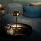 The polished gold Theta Pro Cordless Lamp from Zafferano on an end table next to a couch within a business lounge.