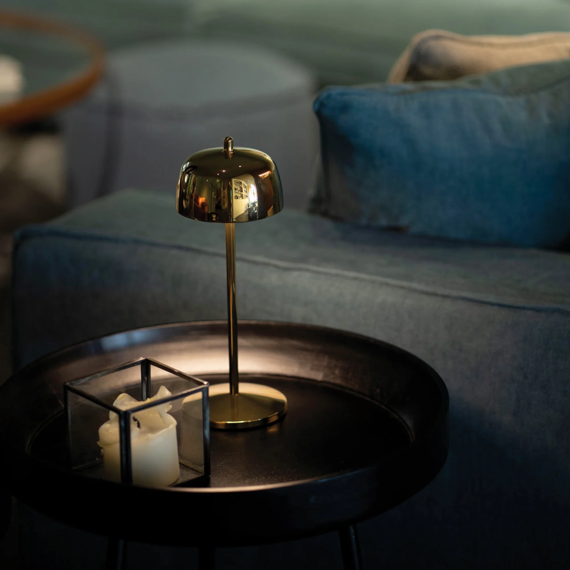 The polished gold Theta Pro Cordless Lamp from Zafferano on an end table next to a couch within a business lounge.