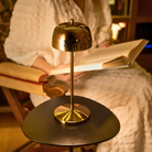 The polished gold Theta Pro Cordless Lamp from Zafferano on top of an end table next to a seat in a reading corner.