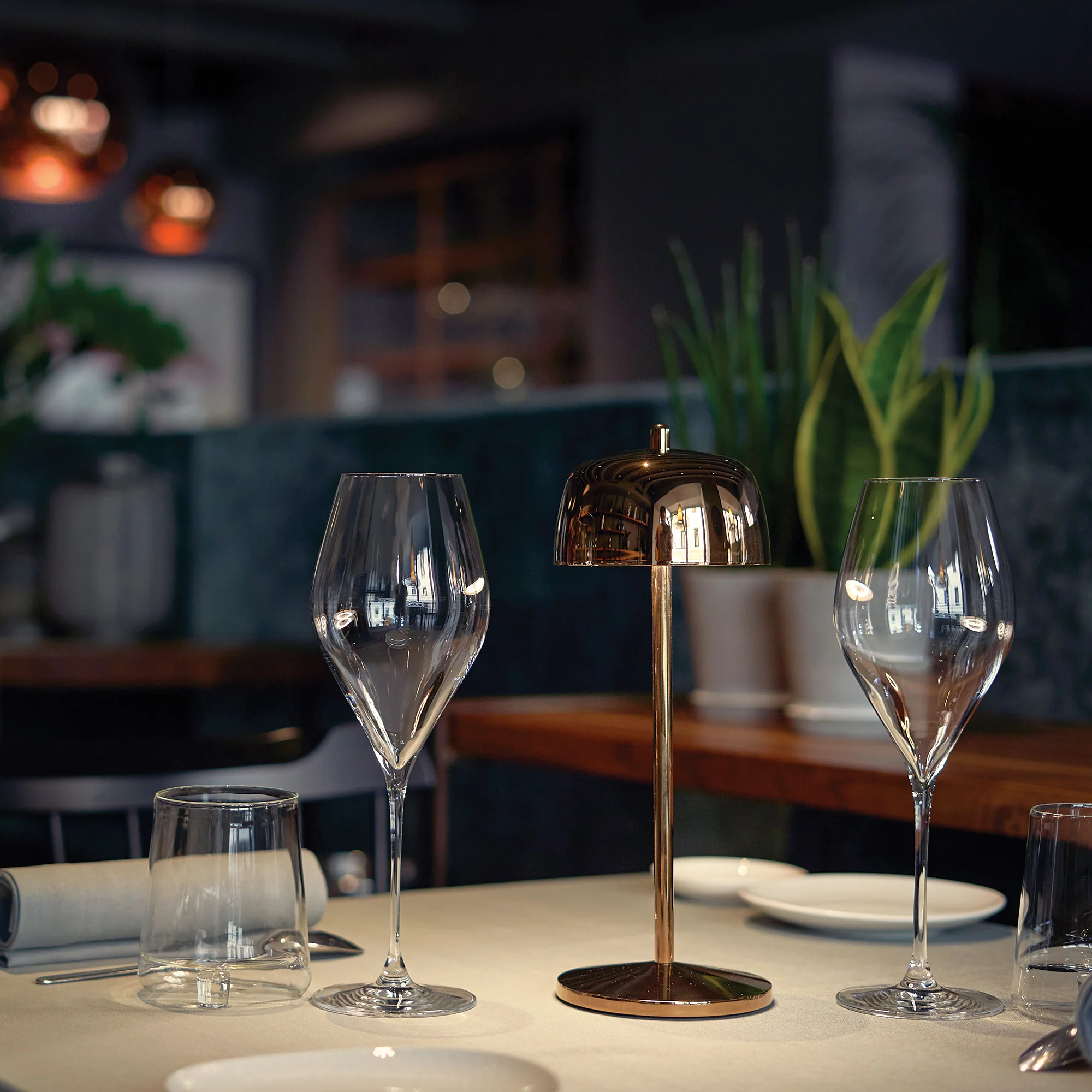 The rose gold Theta Pro Cordless Lamp from Zafferano on the table of a high end restaurant next to wine glasses.