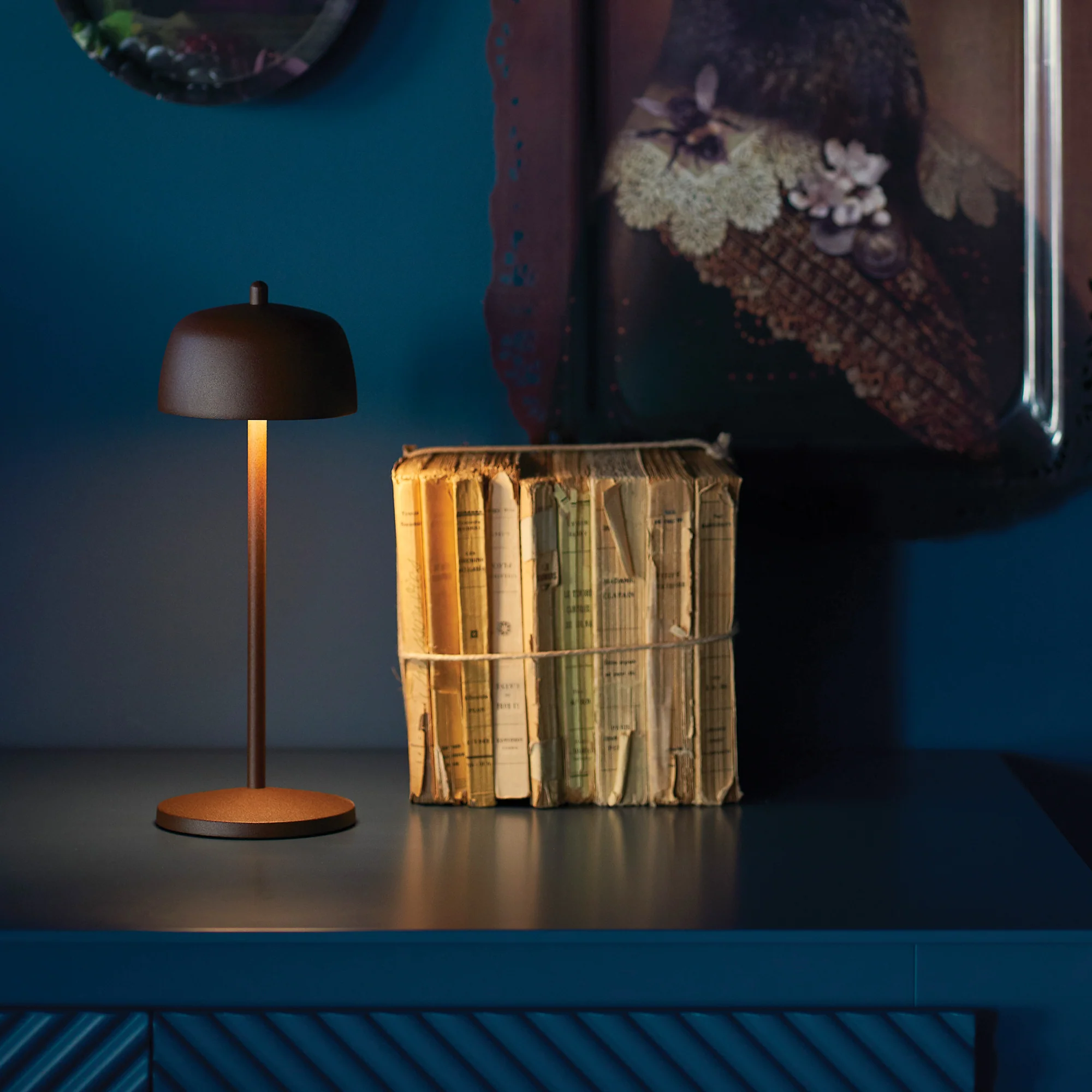 The rust Theta Pro Cordless Lamp from Zafferano in a lounge next to decor on top of a console.